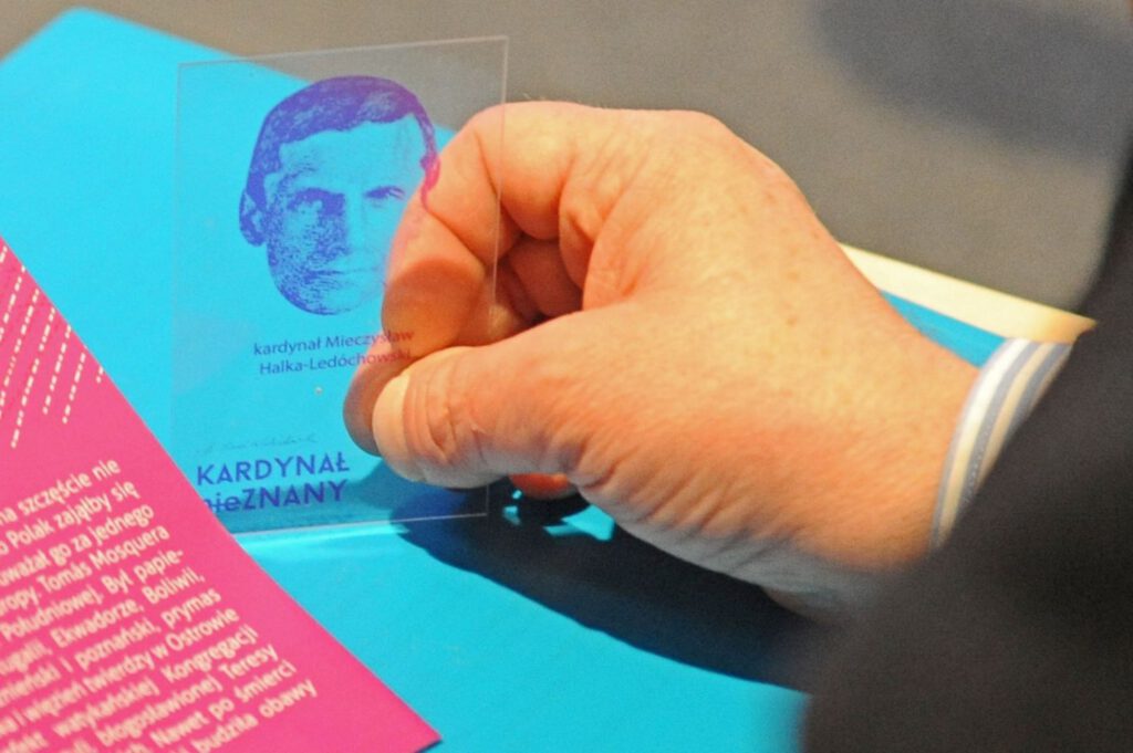 Dłoń trzymająca mała przezroczystą plakietkę z wizerunkiem bohatera wystawy