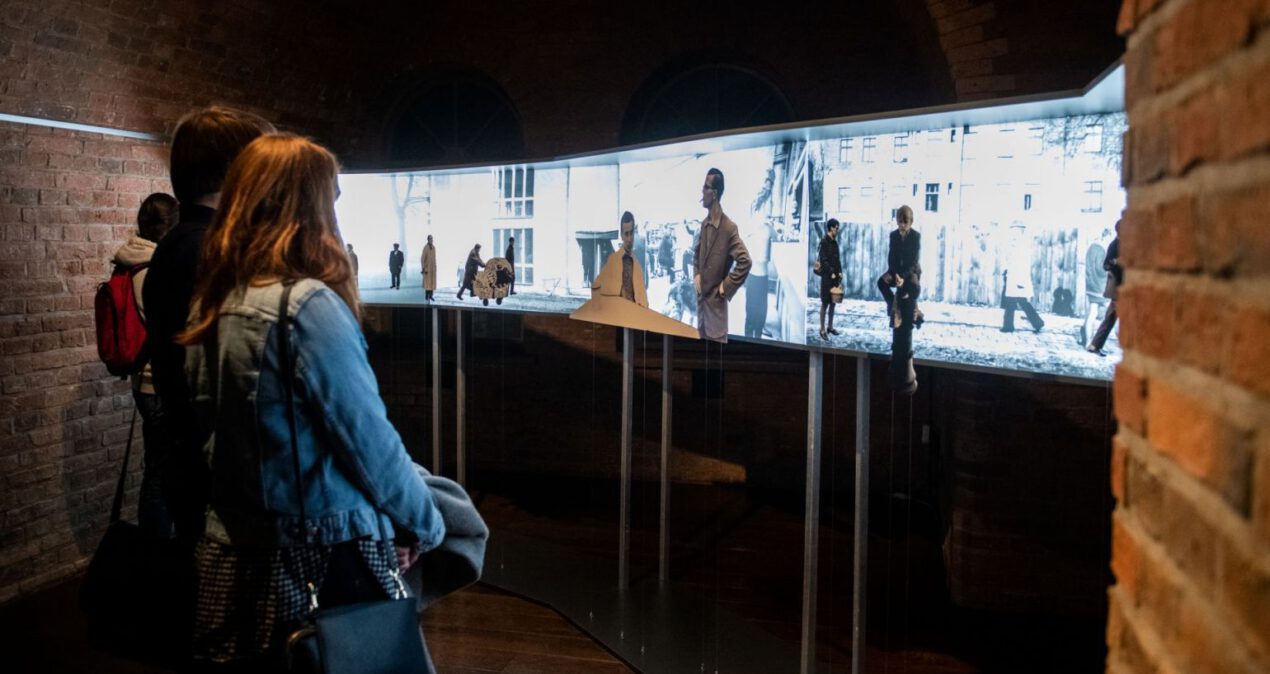 Dziewczyna przygląda się instalacji z podświetlonymi zdjęciami pejzażowymi na których tle umieszczone są różne postaci