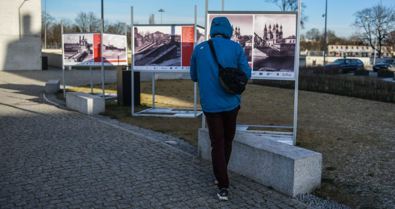 Osoba w niebieskiej kurtce ogląda zdjęcia na palnszy stojącej przed budynkiem Bramy Poznania