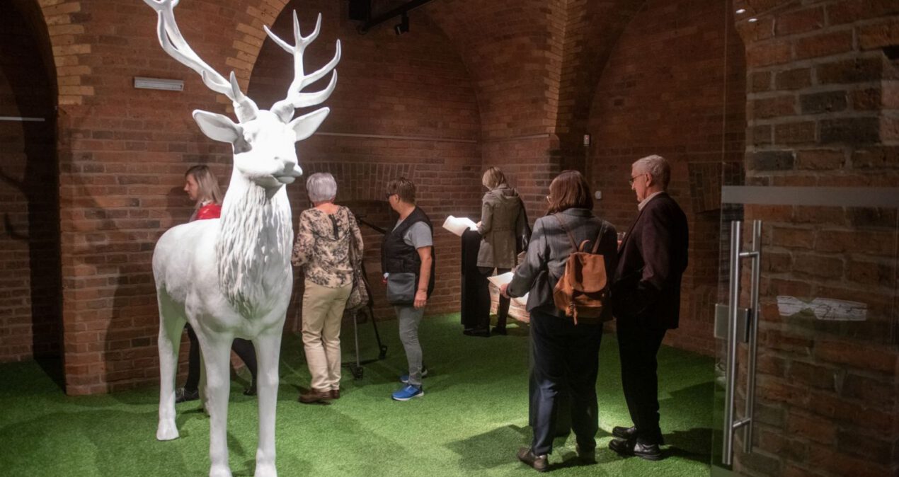Sala wystawowa ze stojącą pośrodku białą figurą jelenia naturalnych rozmiarów. W głębi zwiedzający czytają treści wystawy