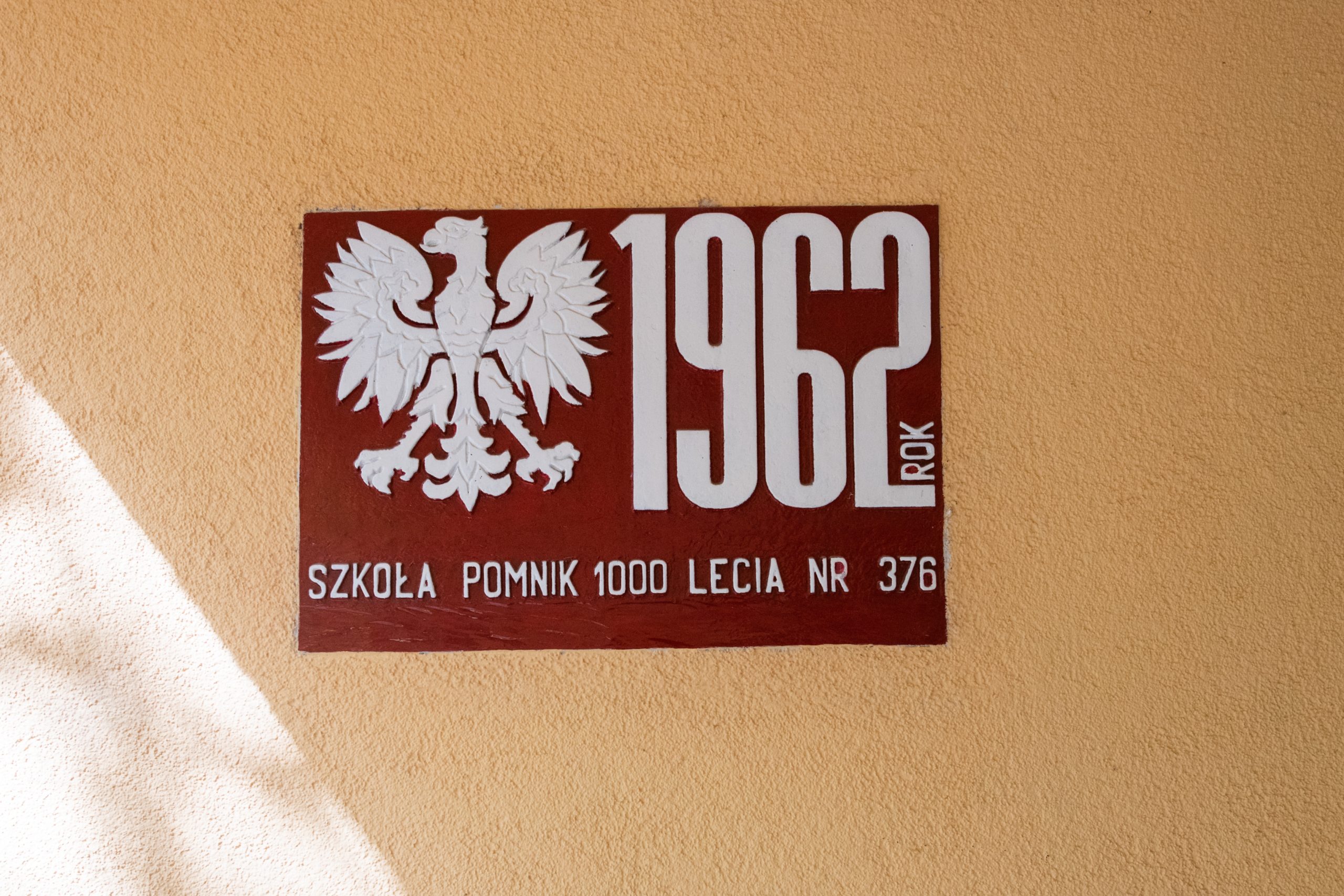 Czerwona tablica przymocowana do żółtej ściany szkoły, na niej napis: 1962 rok Szkoła pomnik 1000-lecia nr 376
