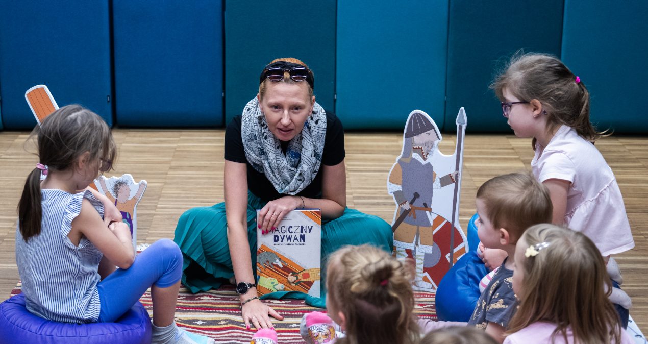 Siedzące na dywanie dzieci słuchają kobiety czytającej książkę