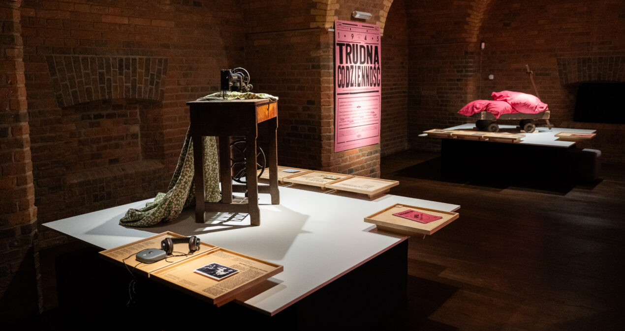 Sala wystawowa, w której znajdują się dwa białe blaty, na których centralnie znajdują się rekwizyty: na jednym stara maszyna do szycia, na drugim stary wózek transportowy na którym leżą dwie poduchy. Wokół nich przymocowane do blatu są drewniane tablice z tekstami wystawy.