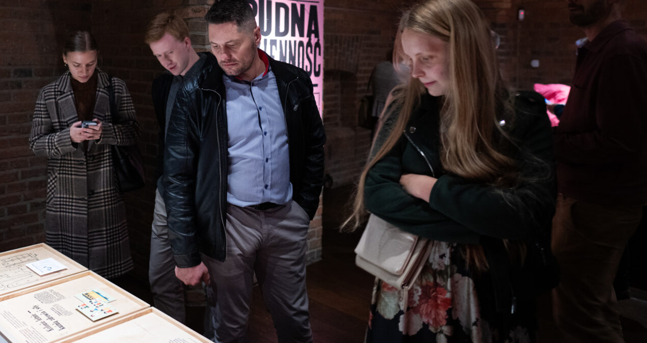 Grupa osób przyglądająca się treściom wystawy umieszczonym na drewnianych tablicach przymocowanych do poziomego blatu