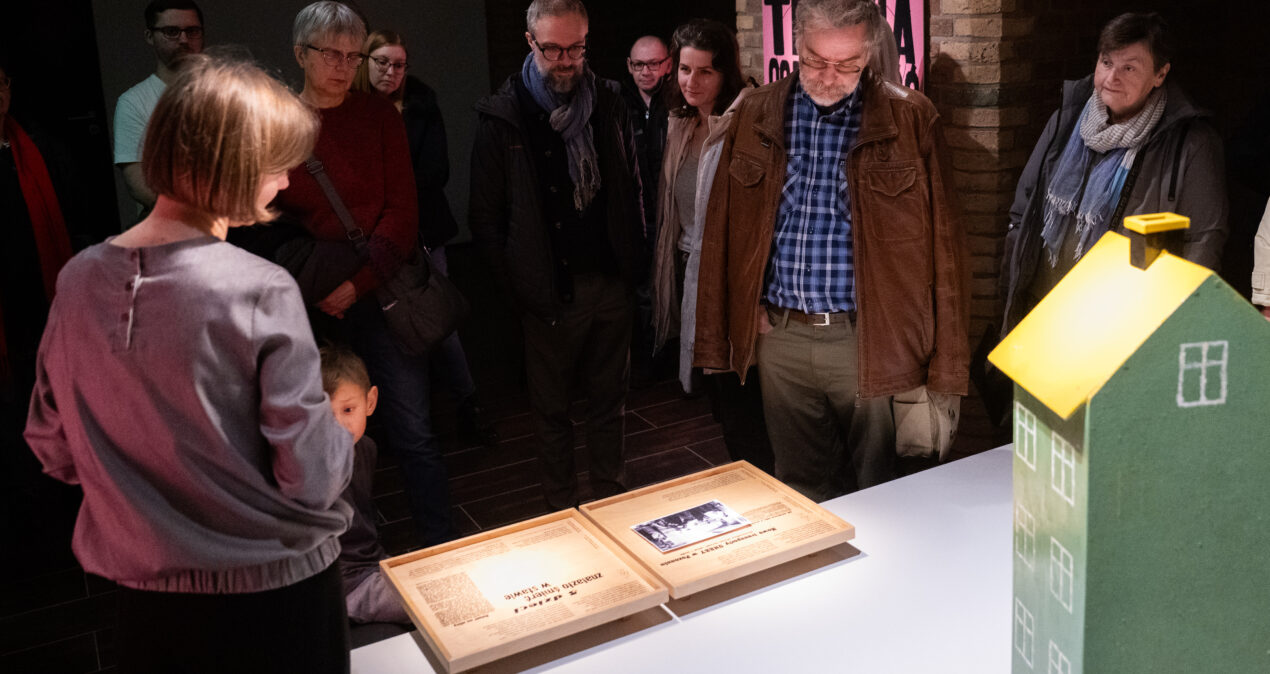 Grupa osób przyglądająca się treściom wystawy umieszczonym na drewnianych tablicach przymocowanych do poziomego blatu, obok których stoi zielony domek do zabawy.