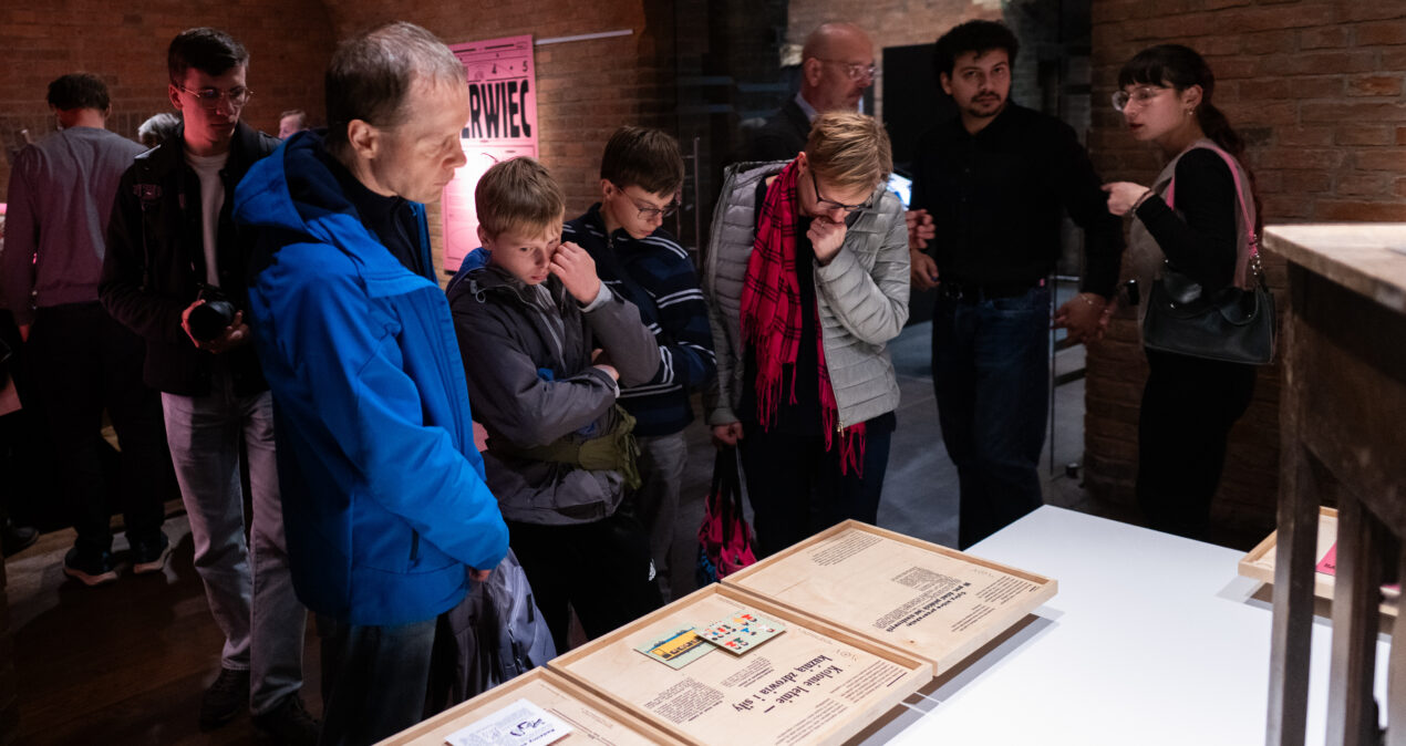 Grupa osób przyglądająca się treściom wystawy umieszczonym na drewnianych tablicach przymocowanych do poziomego blatu, obok których stoi zielony domek do zabawy.