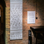 Przestrzeń galerii. Na ceglanej ścianie wisi długie płótno z odbitą fakturą kostki brukowej. To praca Piotra C. Kowalskiego z cyklu obrazy przechodnie